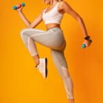 Fitness Lifestyle. Excited Girl Jumping Holding Dumbbells Exercising In Studio Over Yellow Background. Full Length